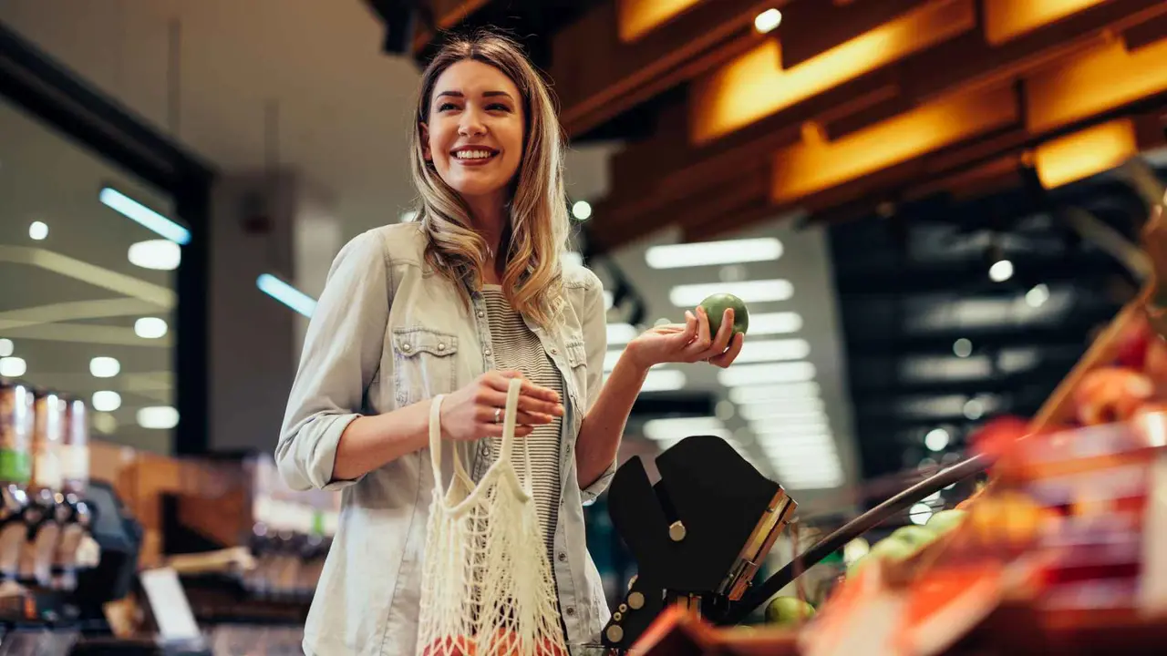 Smiling young woman standing in a grocery store, holding an apple, choosing fresh fruit and putting it in a mesh bag.