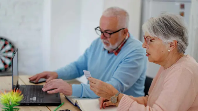 Senior couple uses a laptop and a credit card while sitting at a table in their house.