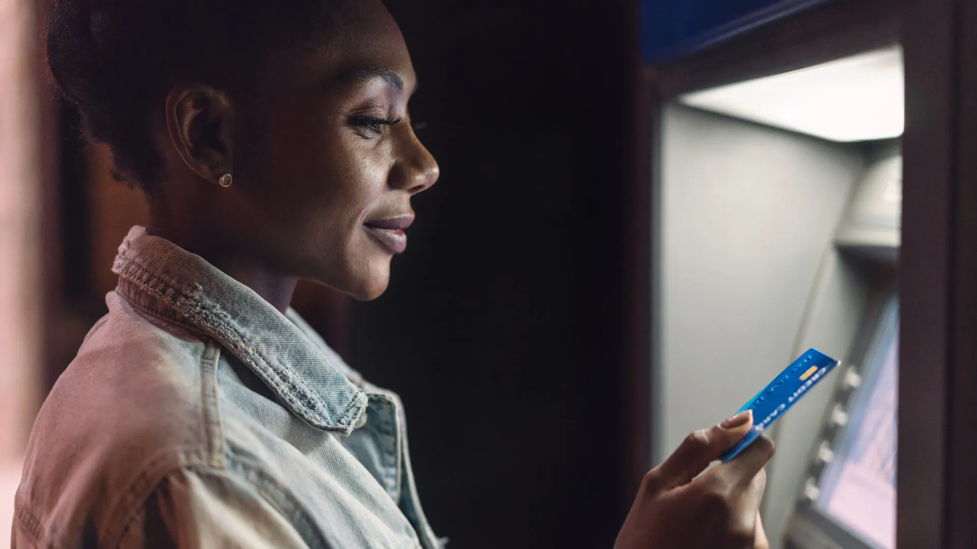 Young African-American woman using her bank card at the ATM in the evening.