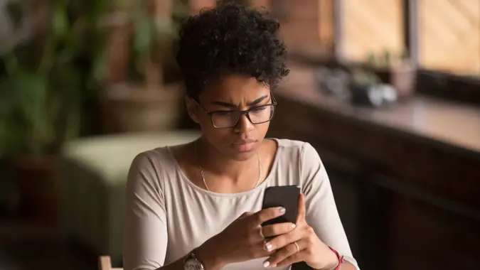 Upset confused african woman holding cellphone having problem with mobile phone, frustrated angry mixed race girl reading bad news in message looking at smartphone annoyed by spam or missed call.