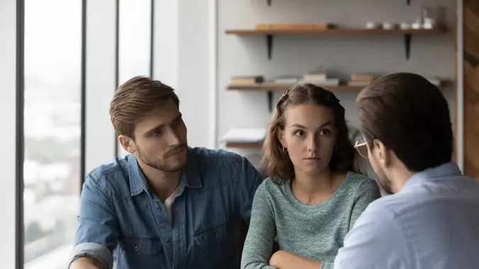 Unhappy young Caucasian couple buyers talk with male Real Estate Agent or real estate agent dissatisfied with contract terms regulations.