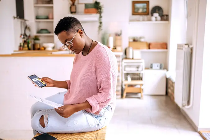 An African-American woman with short hair and glasses, wearing a pink sweater, reviews financial documents in her kitchen.