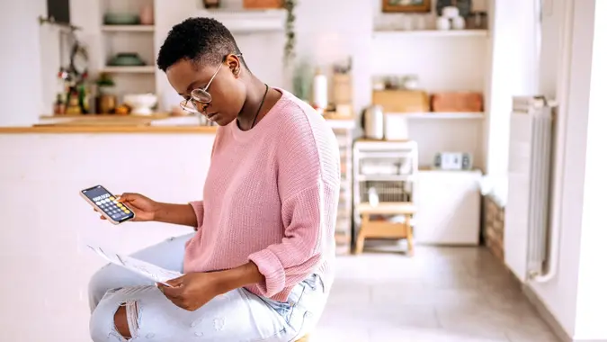 Young woman sitting in her kitchen using a calculator to go over her finances and monthly budget.