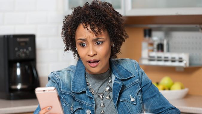 Young African American woman on mobile phone with shocked look on her face, sitting in kitchen.