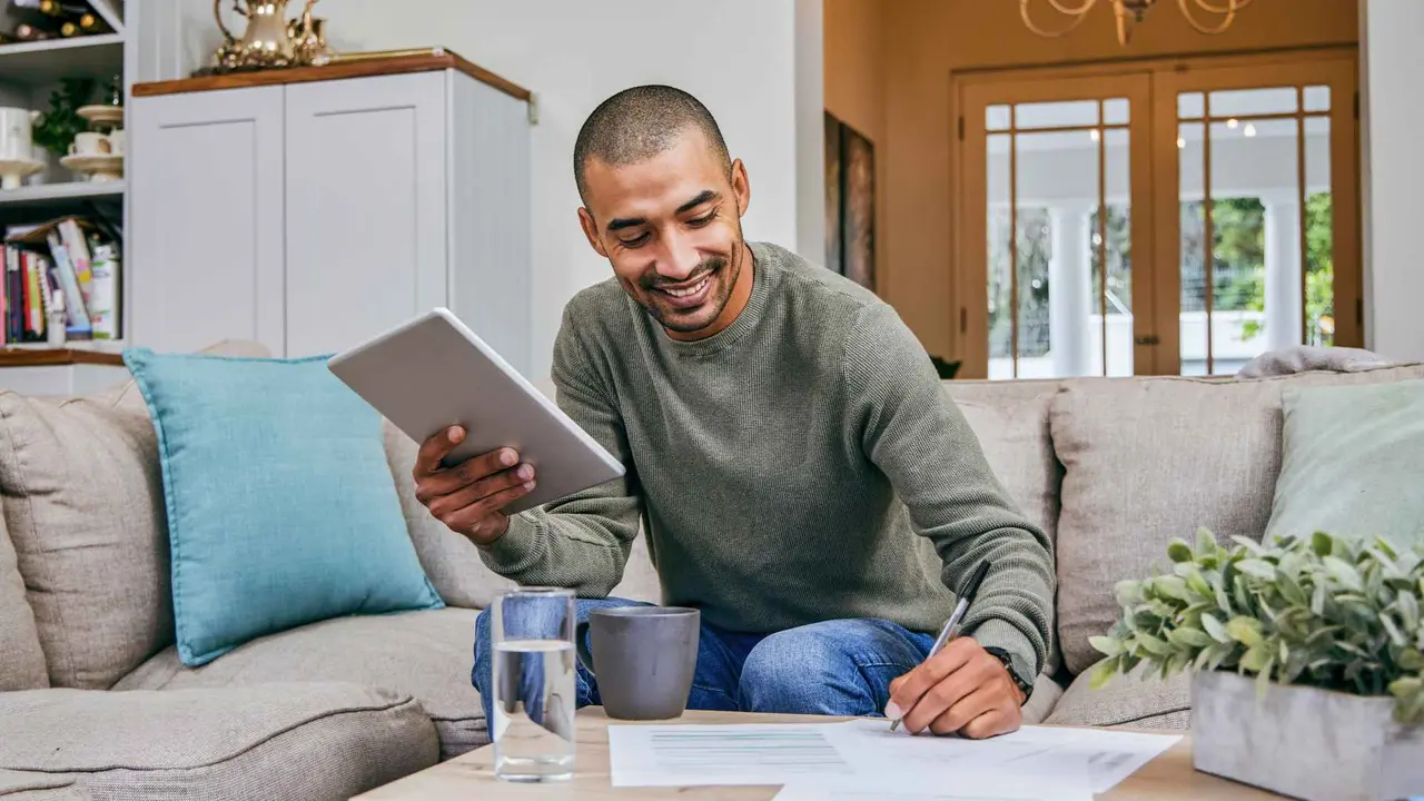 A happy man sitting on his beige living room couch is taking notes while holding his electronic tablet.