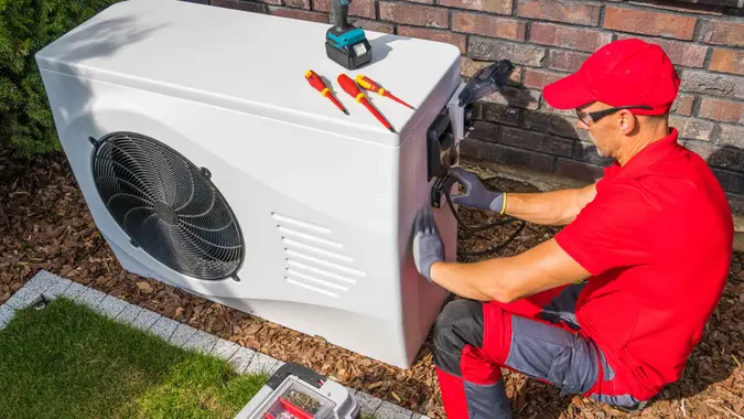 Professional Middle Aged HVAC Technician in Red Uniform Repairing Modern Heat Pump Unit.