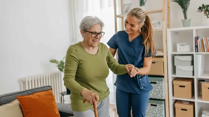 Female caregiver helping and supporting senior patient to walk at home, senior women is standing and holding walking cane.