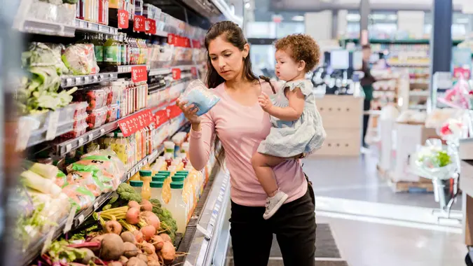 The young mother holds her daughter on her hip as she grocery shops for items she needs.