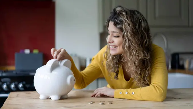 Cheerful young woman with curly hair at home saving coins into her piggybank smiling.