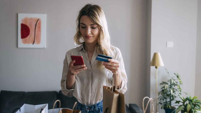 Young female making credit card purchase on her smartphone while standing with few shopping bags on table in living room.