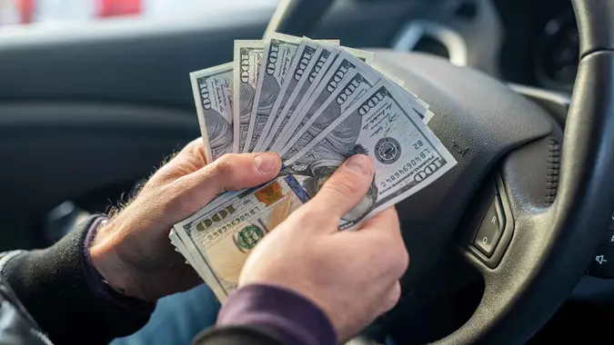 male strong hands holding dollar bills in the middle in the car interior on the background of the steering wheel.