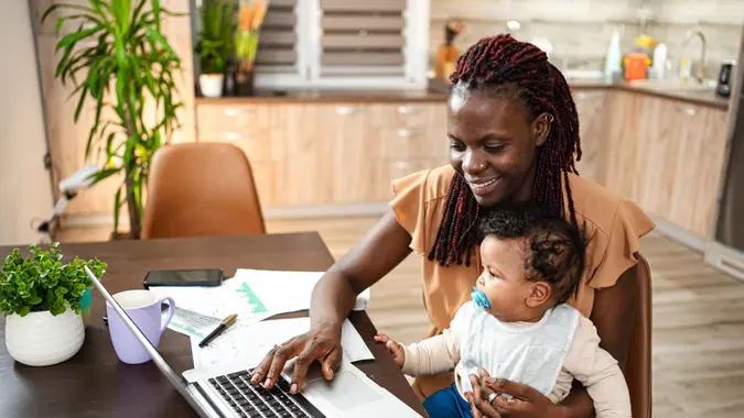 Mother at home reviews finances on the laptop with baby in her lap.