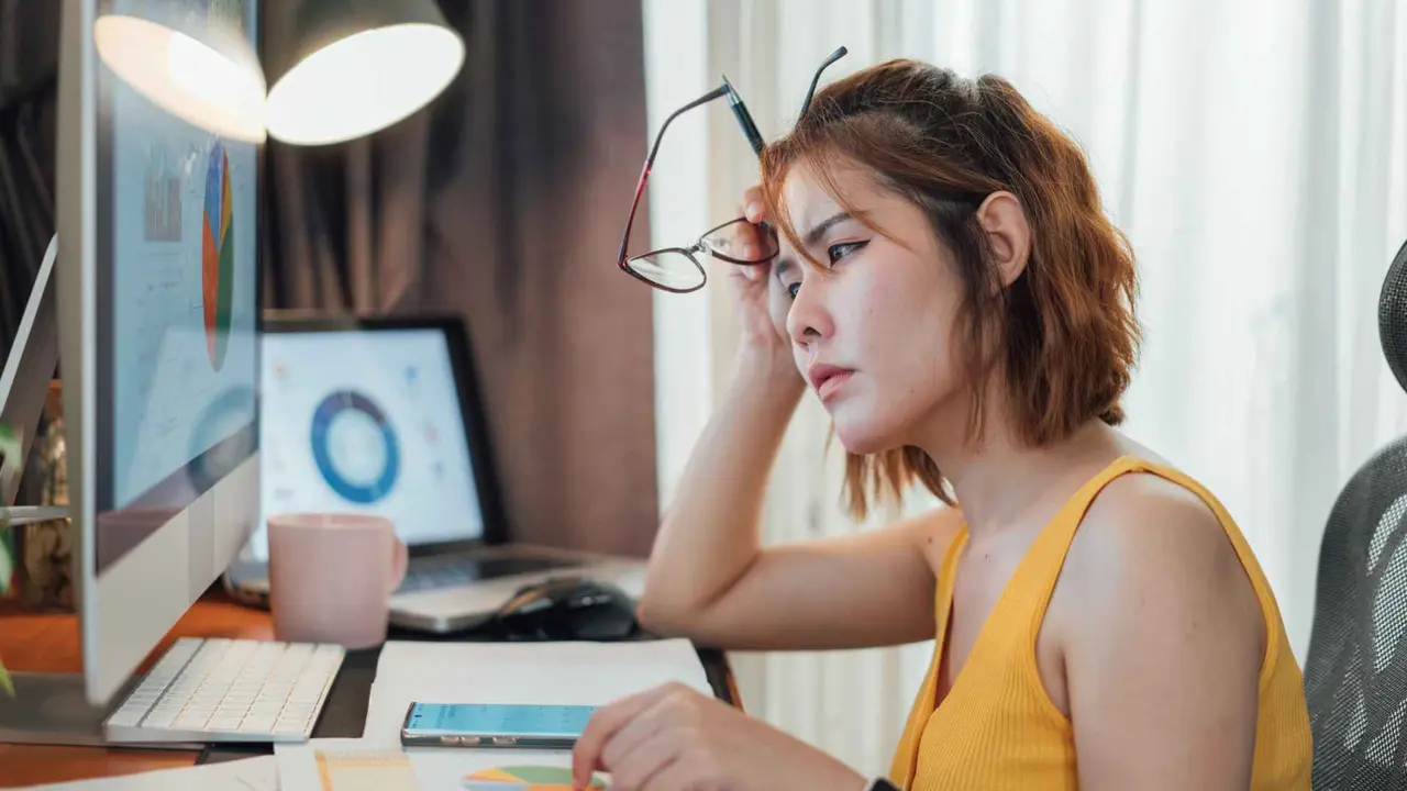 Stressed woman sitting at her desk, looking at a computer with financial charts on the screen.