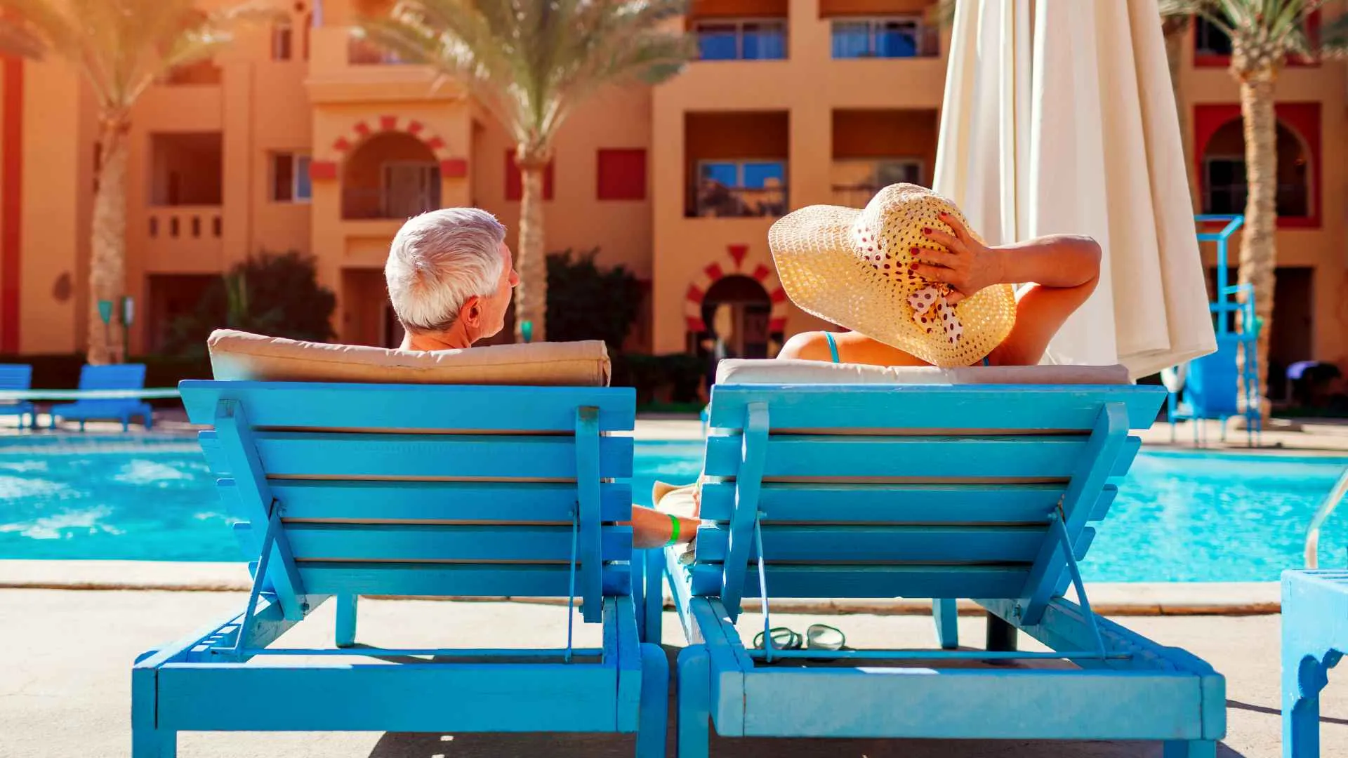A senior couple relaxes while sitting by the pool in their blue lounge chairs.