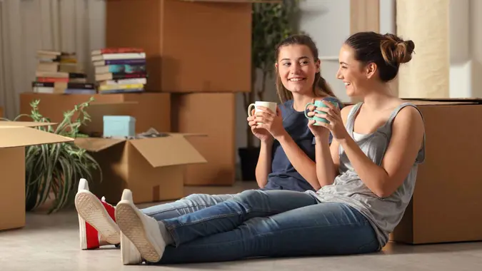 Two happy roommates smile as they sit in their new apartment with boxes.