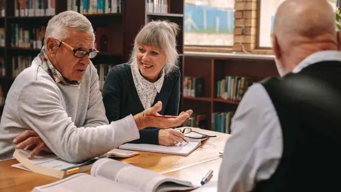 Two senior men and a woman sitting in a library with course books on table and discussing.