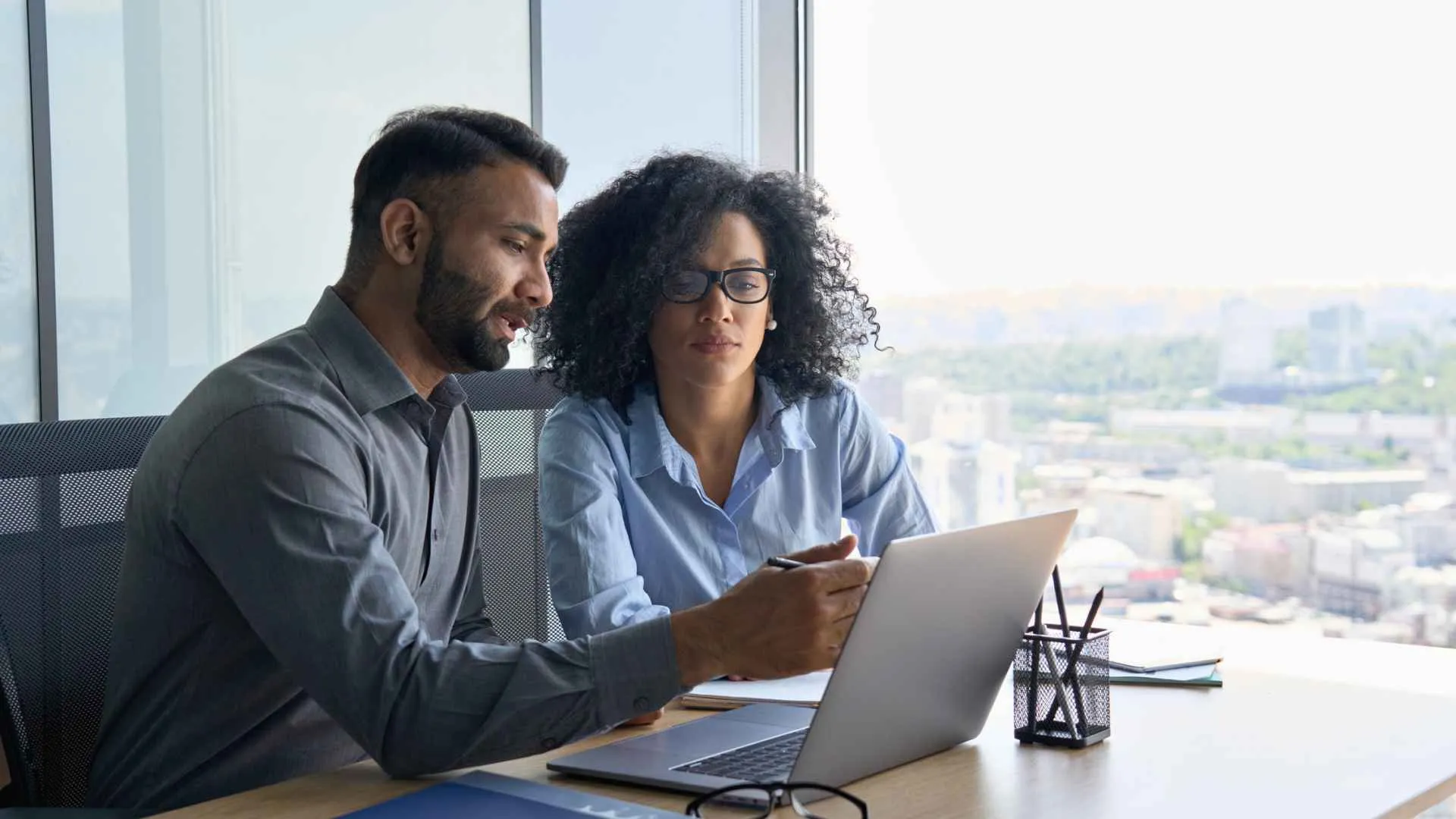 Indian male ceo executive manager mentor giving consultation on financial operations to female African American colleague intern using laptop sitting in modern office near panoramic window.
