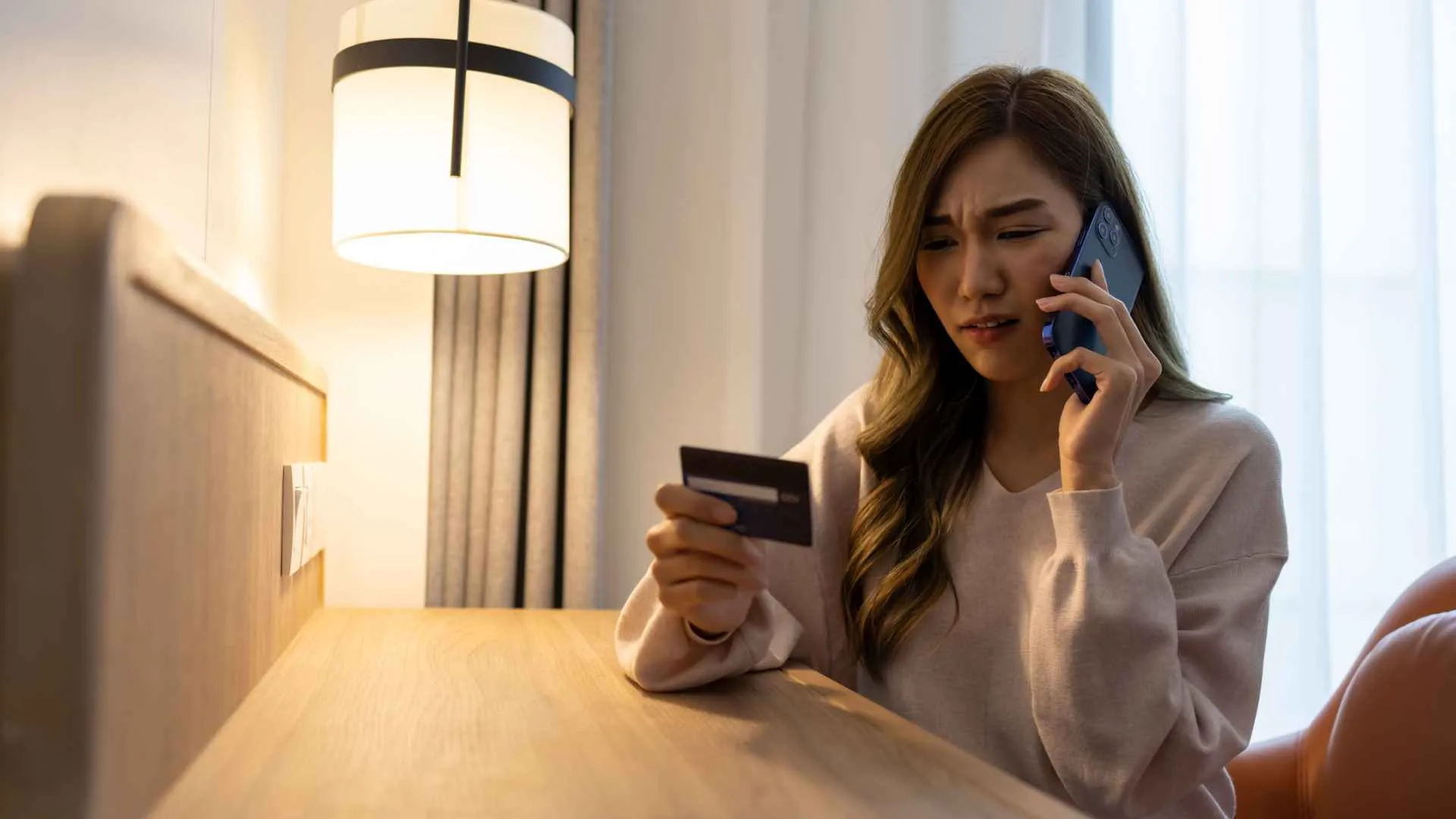 Concerned Asian woman talking on mobile phone while holding credit card on hand.