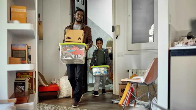 Afro-Caribbean single father in late 20s and 7 year old son smiling as they carry cardboard boxes and plastic containers into new home.