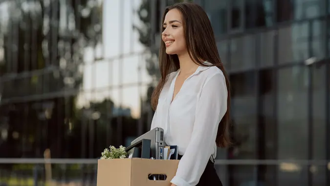 Beautiful smiling brunette girl in a shirt stands in front of the glass modern building where she worked in hands holding a box with packed things, promotion to a position, the joy of quitting stock photo