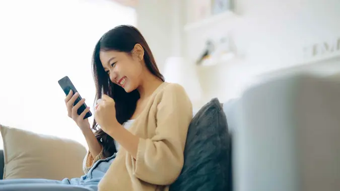 Happy young asian woman relax on comfortable couch at home texting messaging on smartphone, smiling girl use cellphone, chatting online message, shopping online from home.