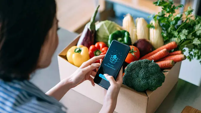 Over the shoulder view of young Asian woman doing home delivery grocery shopping online with mobile app device on smartphone at home, with a box of colourful and fresh organic vegetables and fruits on the table.