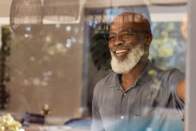 A happy senior man is standing inside looking out through his window at home.