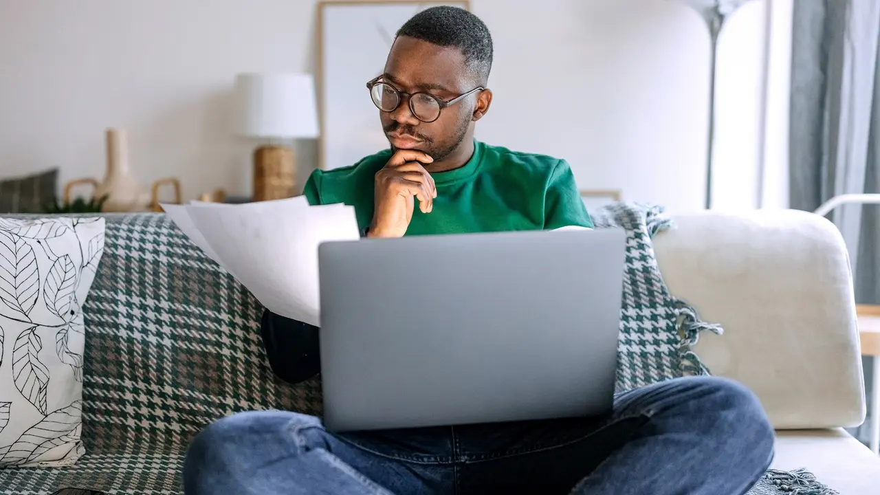 A man sits with his laptop and reads paperwork.