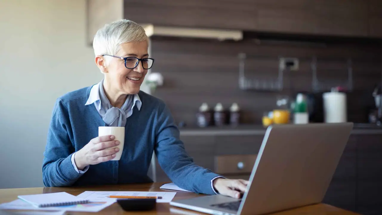 Senior business woman drinking coffee while sitting at her kitchen table using a laptop to go over budget