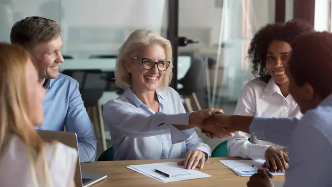 Middle aged woman executive manager company representative welcoming business partner while sitting in modern office boardroom or trainer shake hands with participant before starting education meeting.