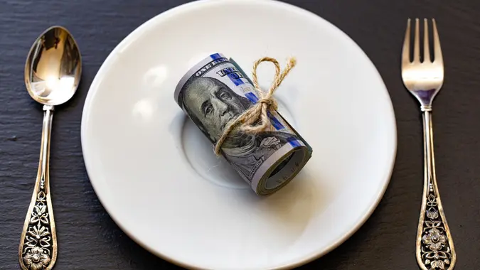 Roll of 100 dollar banknotes on white plate with small fork and spoon stock photo