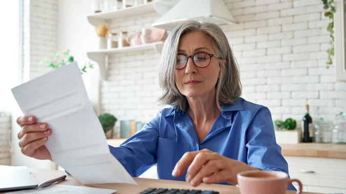 Senior mature business woman holding paper bill using calculator, old lady managing account finance, calculating money budget tax, planning banking loan debt pension payment sit at home kitchen table.