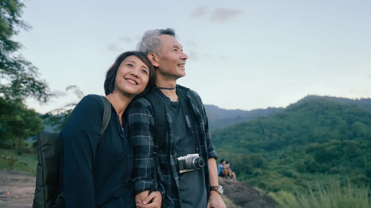 A retired couple smiles while standing outside during a trip.