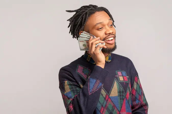 Profile portrait african man holding batch of dollars near his cheek, happy with sudden wealth talking with money.