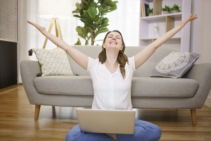 A woman sitting on a sofa with her laptop computer raised her hands in the air, expressing happiness and success.