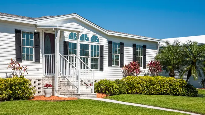 "Mobile home with tropical foliage, shrubbery, palm trees, front lawn, front door, sidewalk.