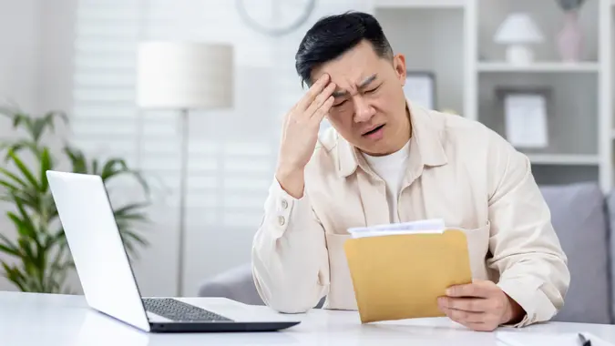 Upset and disappointed man in home office received message mail envelope with bad news, asian man working in home office at desk with laptop.