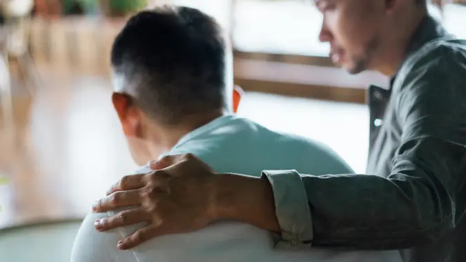 Rear view of son and elderly father sitting together at home. Son caring for his father, putting hand on his shoulder, comforting and consoling him. Family love, bonding, care and confidence stock photo