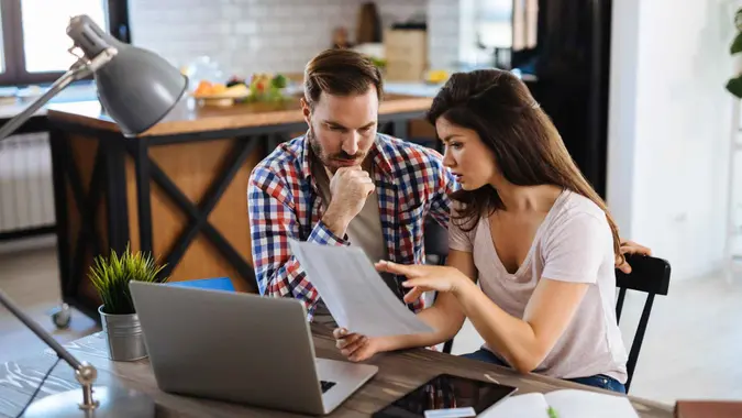 A couple goes over their monthly bills while reading a statement in front of their laptop.