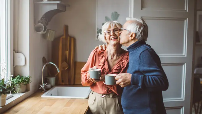 A happy retired couple stands in their kitchen as the husband give his wife a kiss on the cheek.