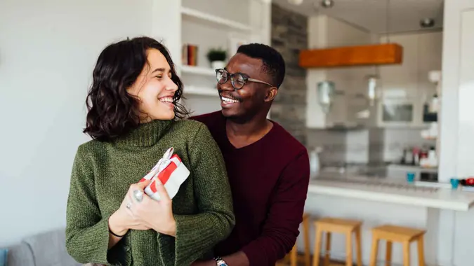 Happy young interracial couple having fun at home, African American boyfriend surprising his Caucasian girlfriend with a Valentine''s day present, making her smile.