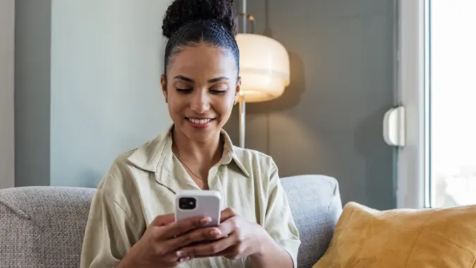 A happy woman looks at her phone while working on her laptop at home.