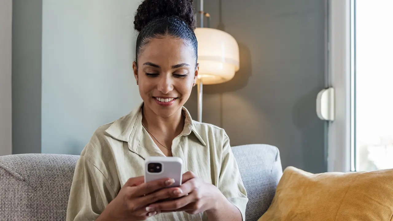 A happy woman looks at her phone while working on her laptop at home.