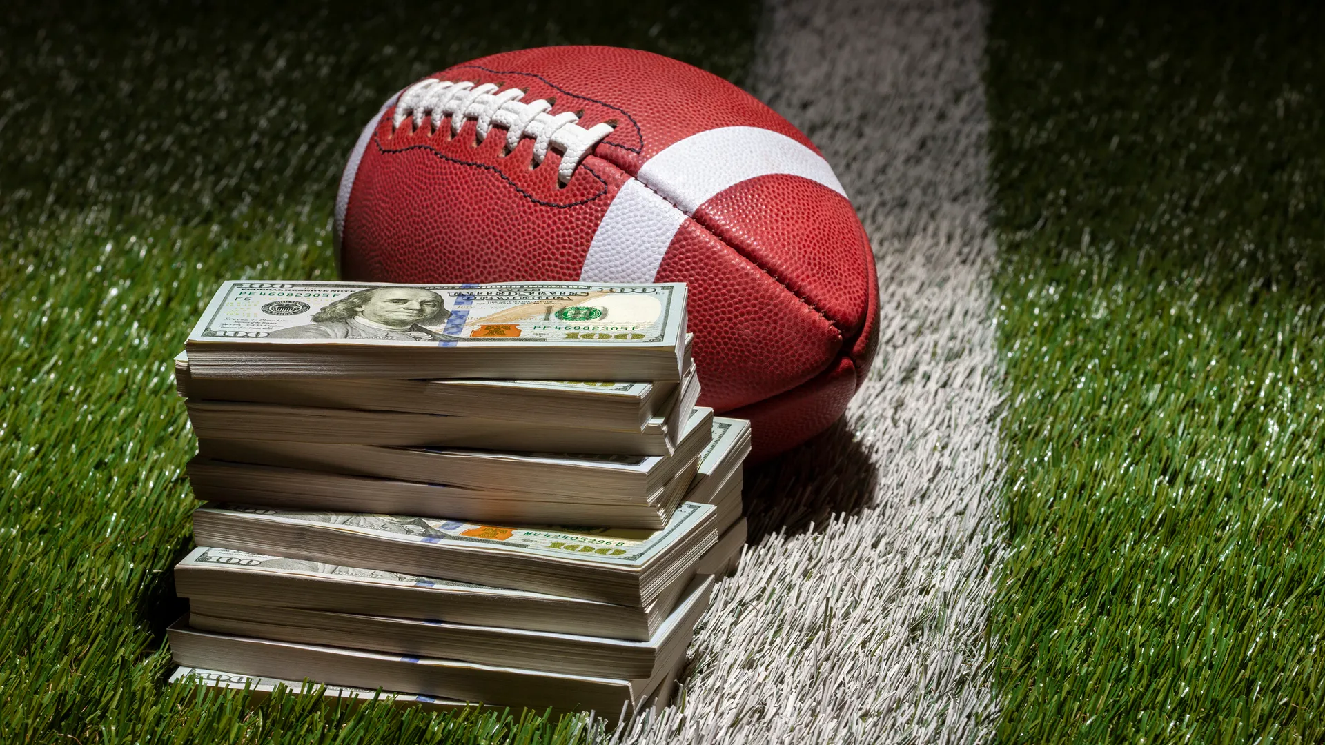 A football and a pile of one hundred dollar bills on a grass field with stripe and dark background.