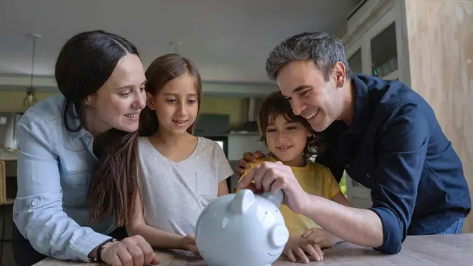 Loving latin american parents teaching their kids to save money in a piggy bank all smiling.