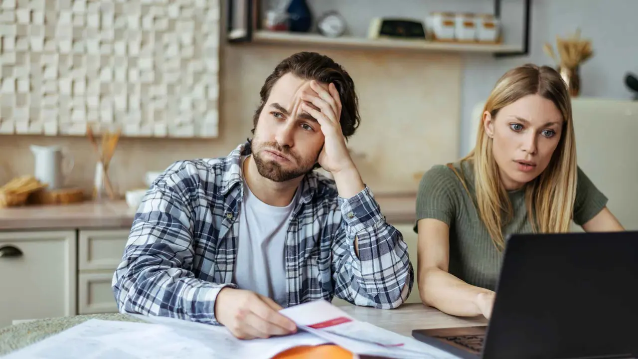 Upset european millennial man with stubble and blonde woman pay bills and taxes with laptop in light kitchen interior.