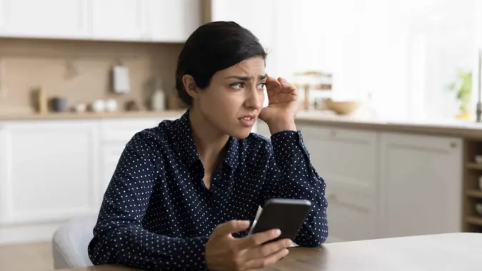 Concerned young Indian woman holding smartphone, looking away in deep thoughts, thinking on problems, feeling doubt, uncertain, worried, anxious, touching head, chatting at home table.