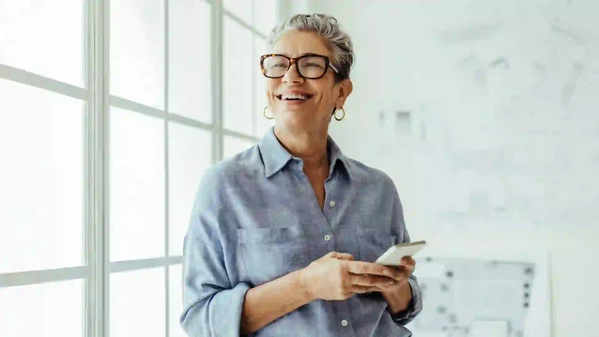 A happy senior woman holds her phone and smiles while standing in front of a window in her home.