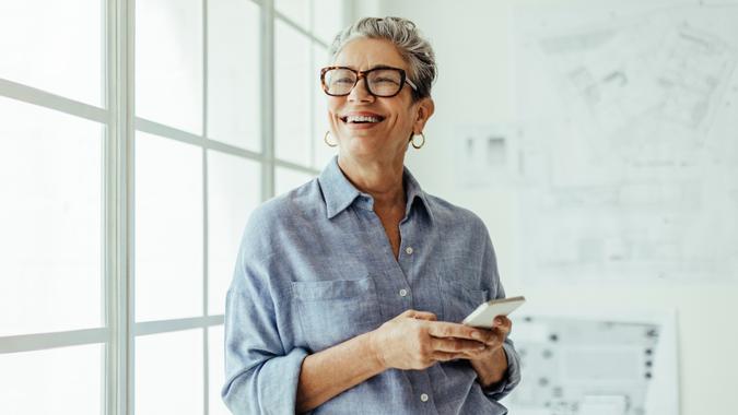 A happy senior holds her phone and smiles while standing in front of the window.