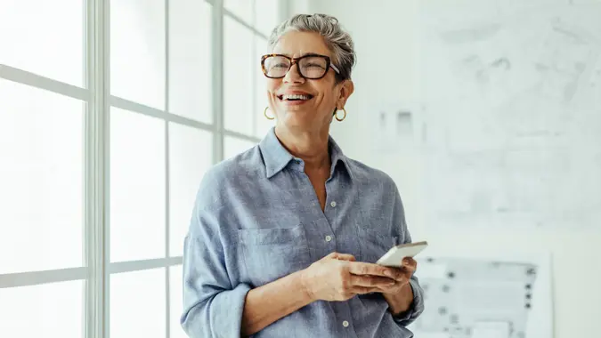 A happy senior holds her phone and smiles while standing in front of the window.